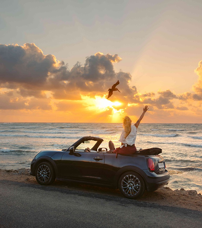 Front left view of the MINI Cooper Convertible in Copper Grey at a beach with two people next to it.